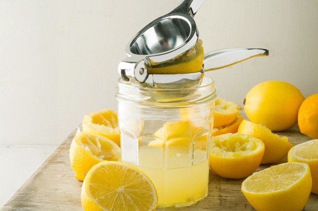 A metal, handheld citrus juicer over a clear glass jar that's half-filled with lemon juice. Around it are squeezed and un-squeezed lemon halves.