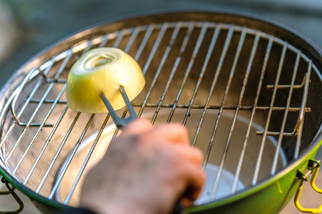 Anonymous person using a halved onion skewered with a grill fork to clean the grates of a round charcoal grill.