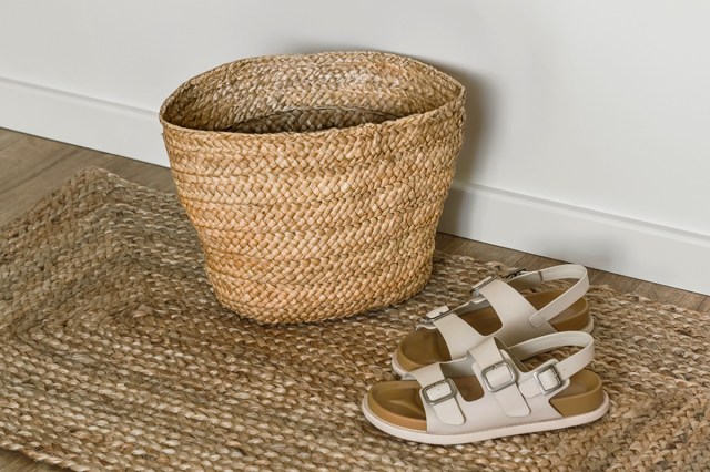 A woven seagrass storage basket placed on a jute rug next to a pair of white leather sandals.